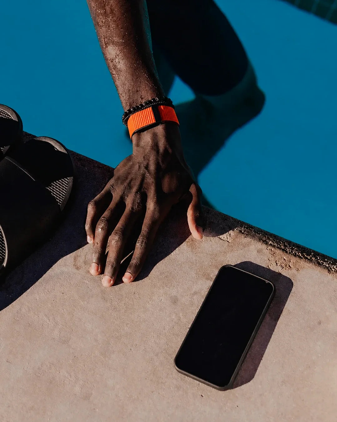 Mão com a pulseira QBand laranja na borda da piscina, com o celular deixado de lado
